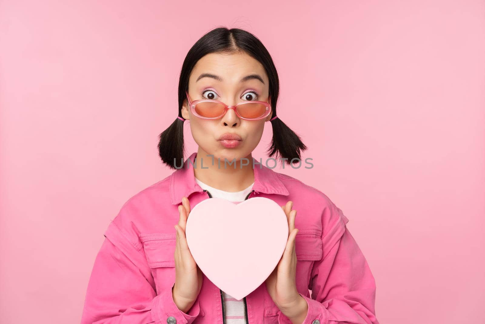 Cute asian girl showing heart shaped box with gift, looking surprised and excited, romantic present concept, wearing sunglasses, standing over pink background.