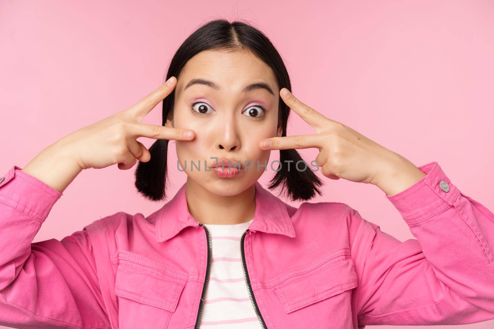 Close up of stylish asian girl smiles happy, shows peace v-sign, kawaii pose, posing against pink background. Copy space