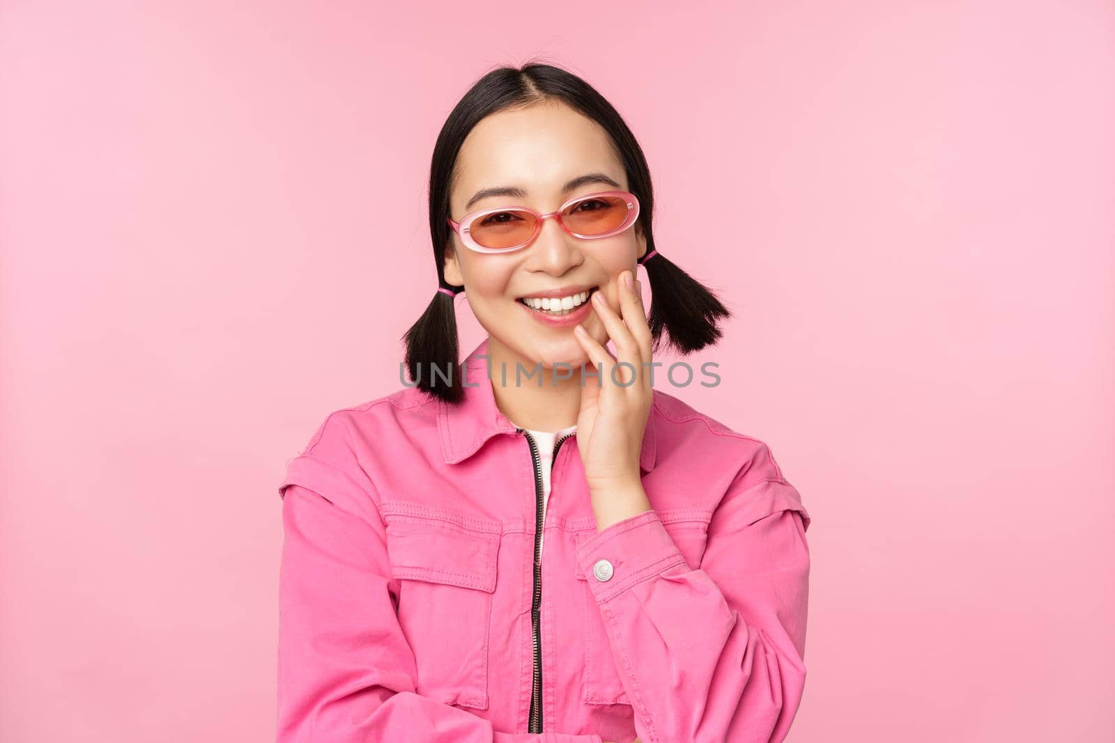 Close up portrait of stylish asian woman in sunglasses, smiling, looking flattered, laughing coquettish, standing over pink background. Copy space