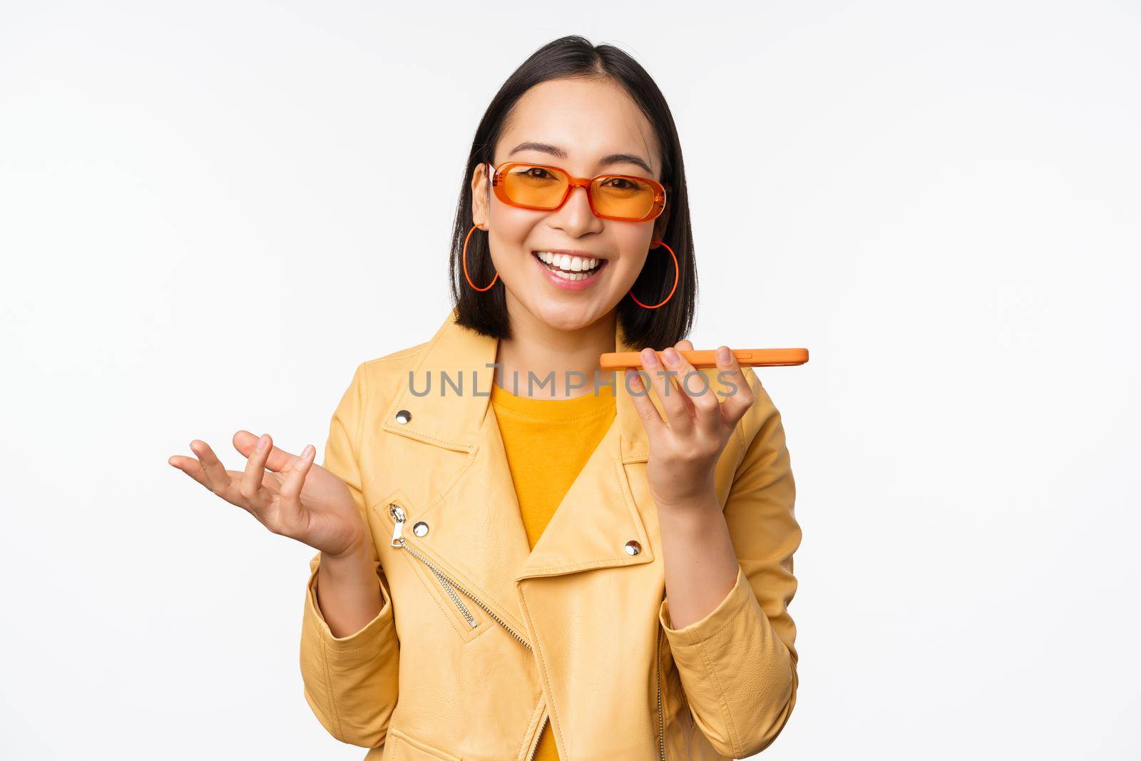 Image of happy asian girl talking on speakerphone, recording, translating her voice with mobile phone app, talking in smartphone dynamic, standing over white background.
