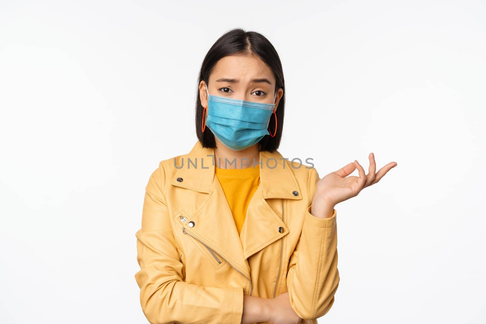 Confused asian woman in medical face mask, looking clueless, puzzled, wearing medical face mask, standing over white studio background by Benzoix