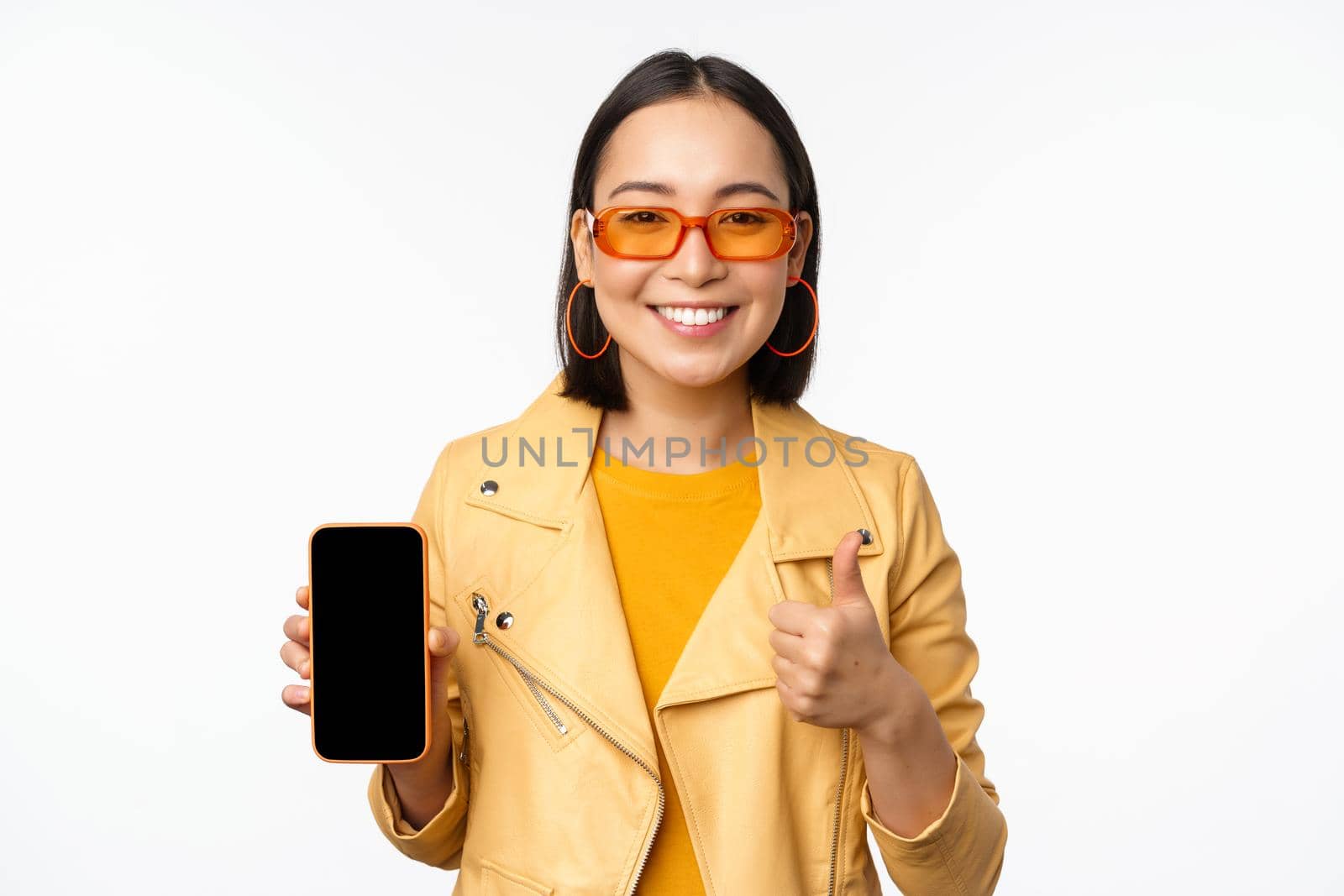 Beautiful korean girl, asian woman in sunglasses, showing smartphone app interface, thumbs up, recommending mobile phone application, white background by Benzoix