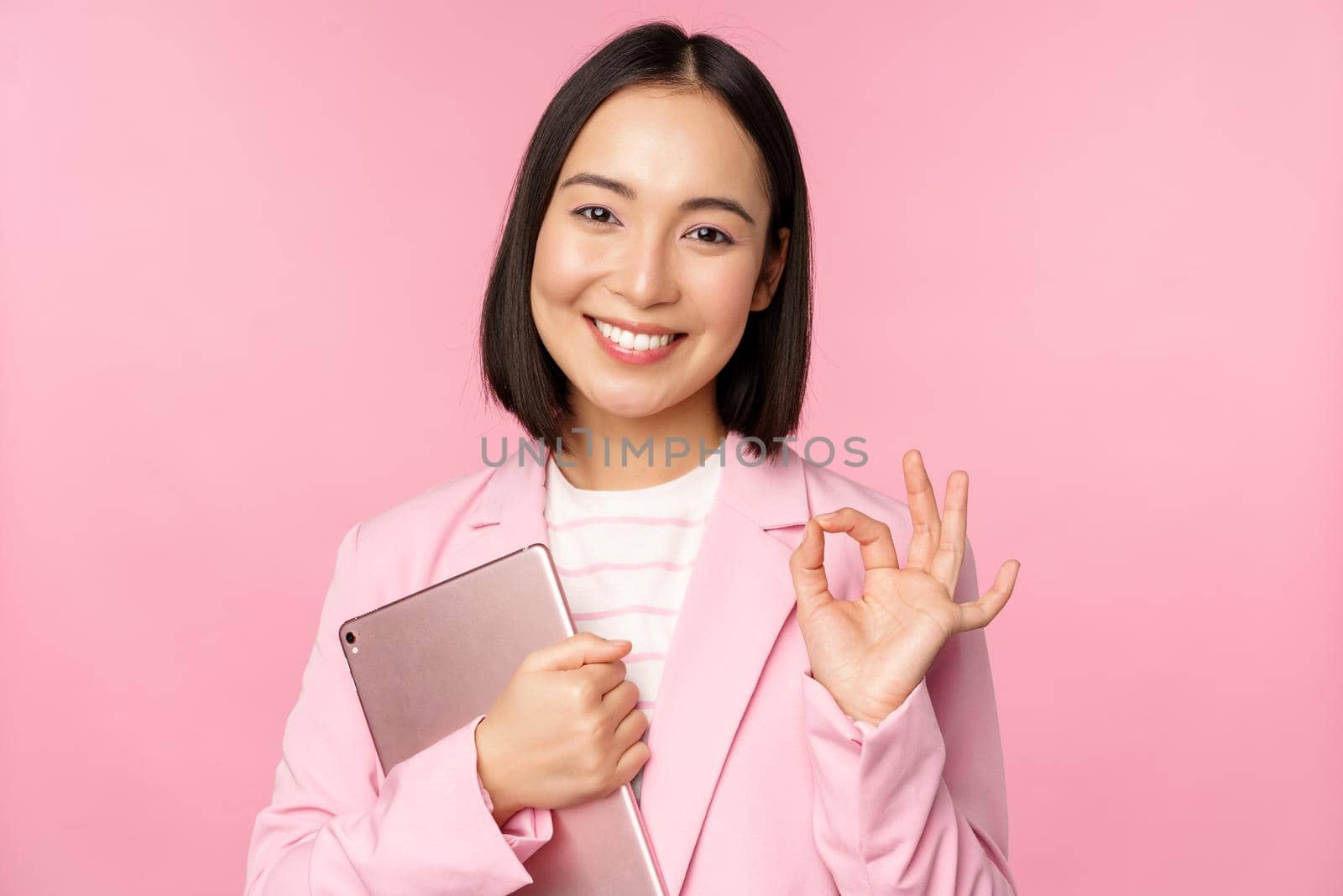 Portrait of corporate woman, girl in office in business suit, holding digital tablet, showing okay, recommending company, standing over pink background.