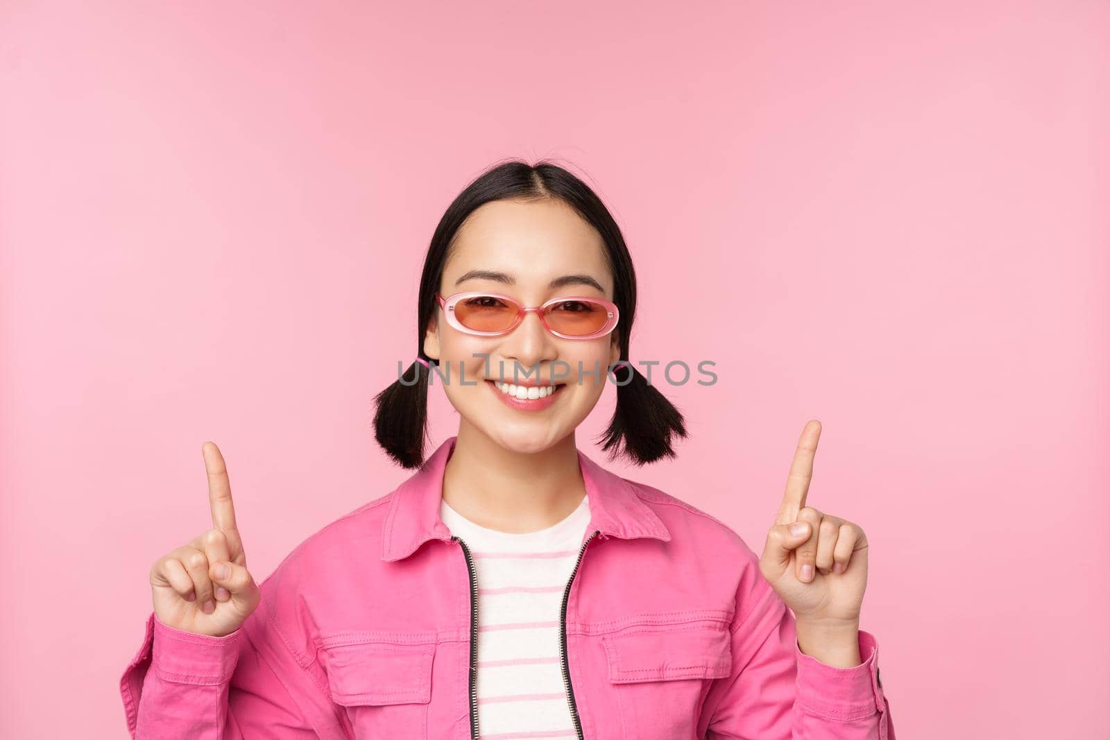 Close up portrait of modern korean female model, wears sunglasses, points fingers up, shows advertisement, promo banner, pink background. Copy space