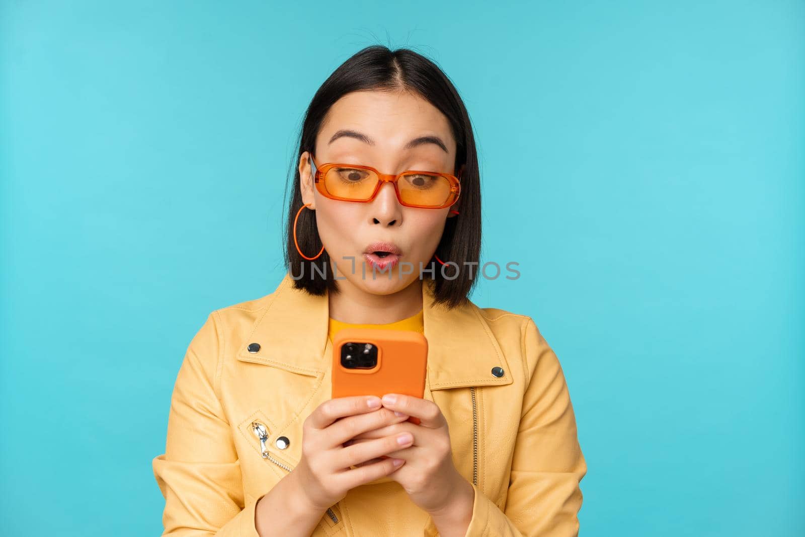 Portrait of asian woman in sunglasses, looking at mobile phone with surprised face expression, standing over blue background. Copy space