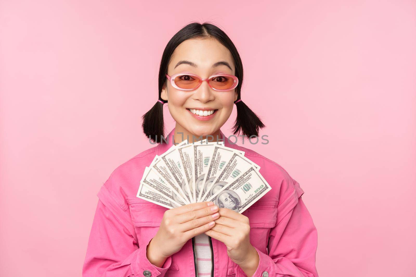 Microcredit and fast loans concept. Excited stylish korean girl, showing money, cash dollars and looking happy, standing in sunglasses over pink background by Benzoix