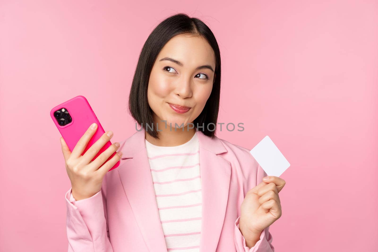 Smiling asian corporate woman, lady in suit thinking, holding smartphone and credit card, plan to buy smth online, shopping with mobile phone, pink background.