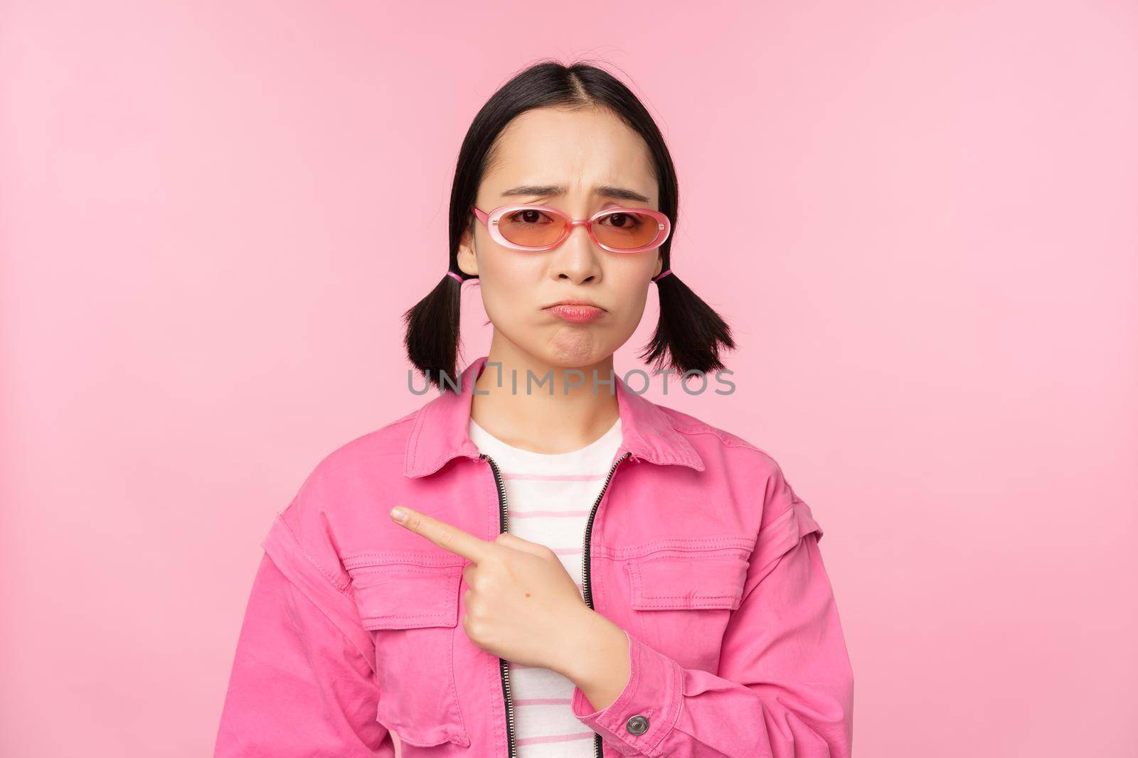 Potrait of cute korean girl in sunglasses, pointing left and looking disappointed, sulking upset, standing over pink background by Benzoix