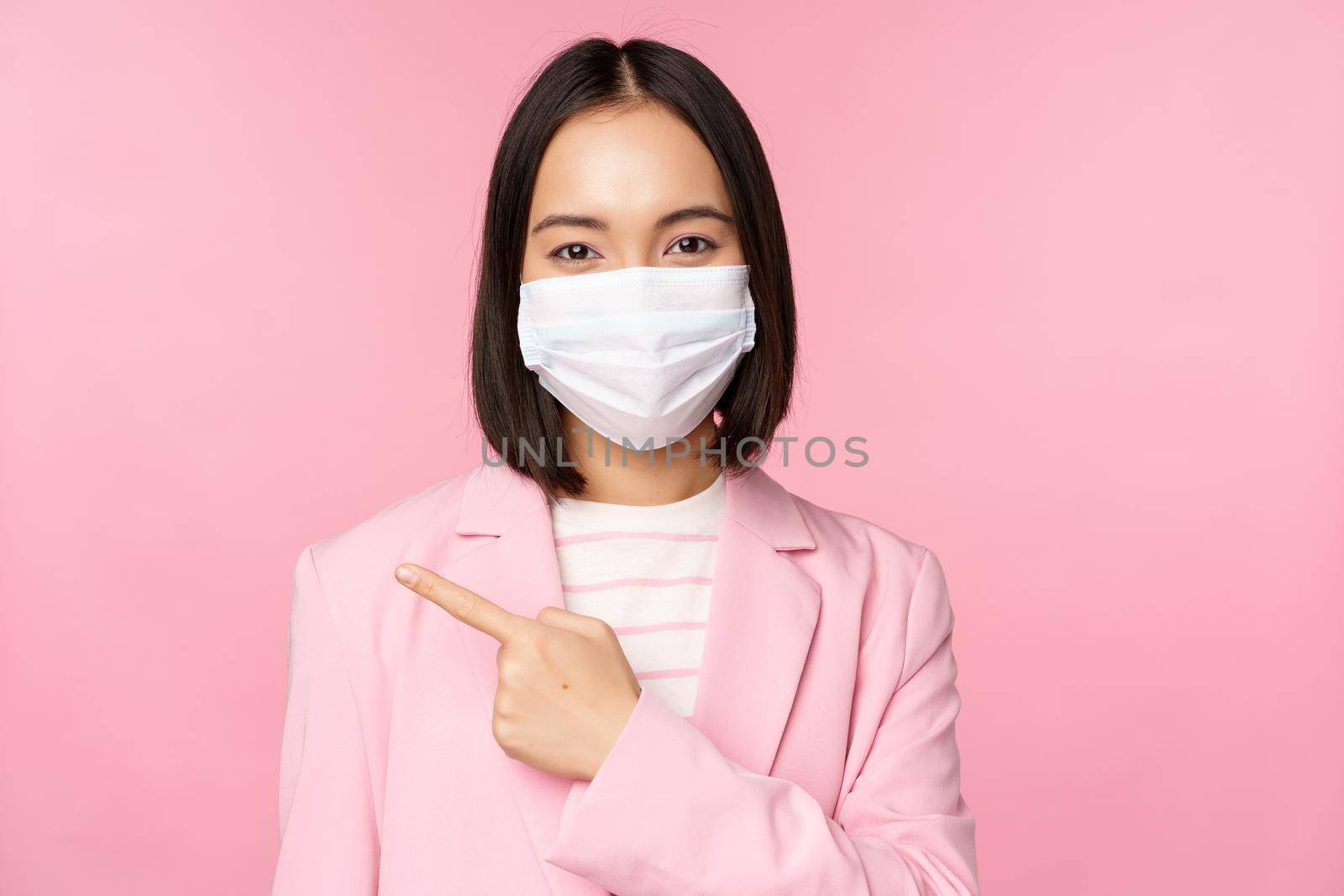 Portrait of asian businesswoman in medical face mask and suit, pointing finger left, showing advertisement, company banner, studio pink background.