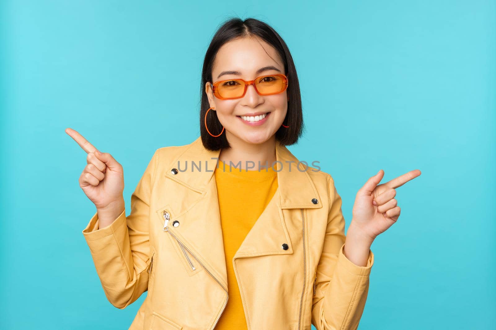 Happy stylish chinese girl in sunglasses, points fingers sideways, invites to check out, see store discount, shows left and right, stands over blue background by Benzoix