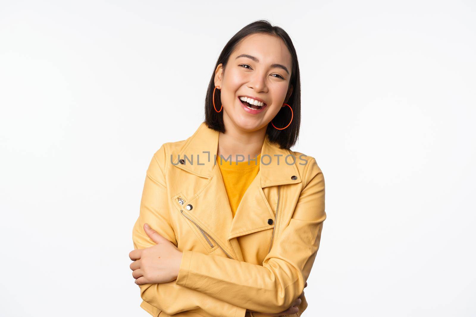 Image of beautiful modern asian girl laughing, smiling and looking happy at camera, standing in yellow jacket against white background by Benzoix