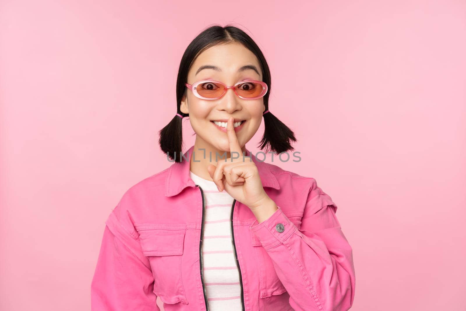 Modern korean girl in stylish spring outfit, sunglasses, showing shush, hush sign, press finger to lips, taboo gesture, standing over pink background by Benzoix