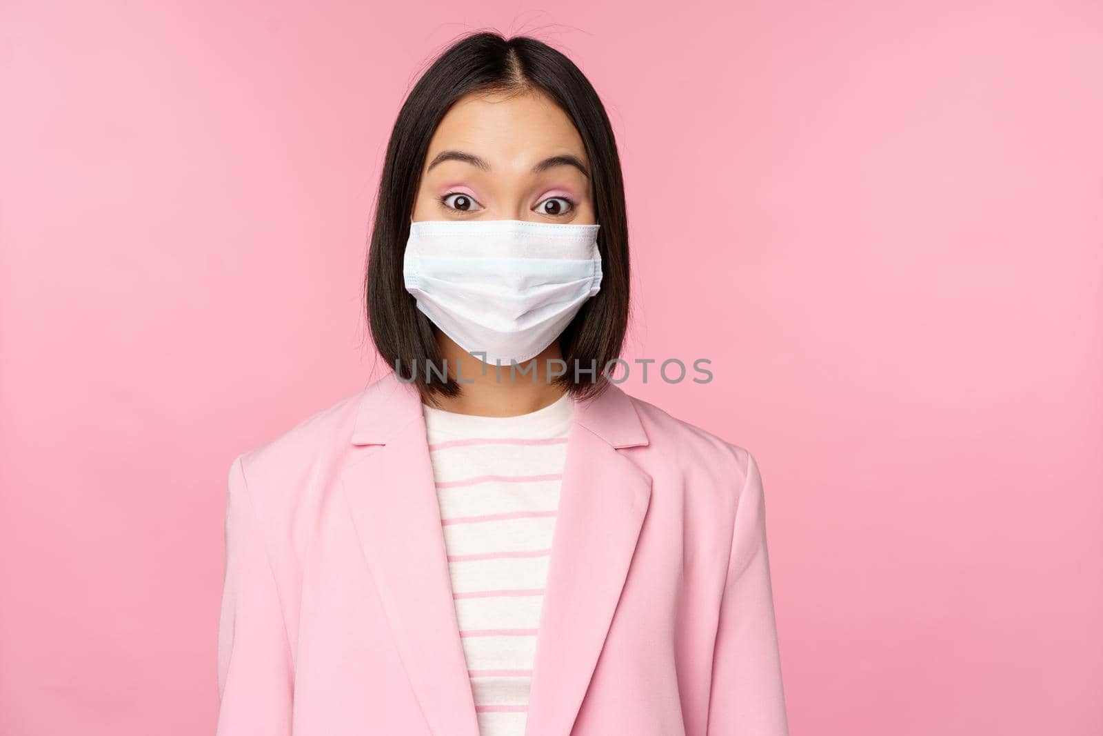 Portrait of asian businesswoman in medical face mask, wearing suit, concept of office work during covid-19 pandemic, standing over pink background.