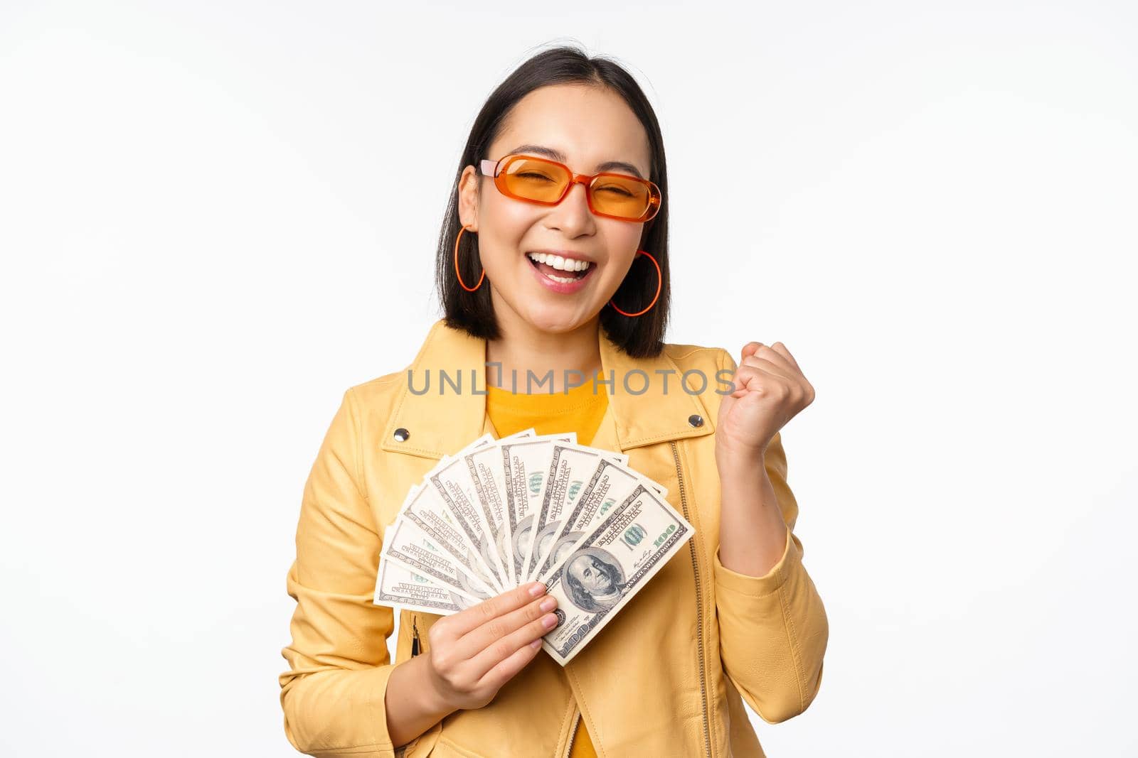 Stylish smiling asian girl holding money cash, showing dollars and celebrating, standing over white background. Copy space