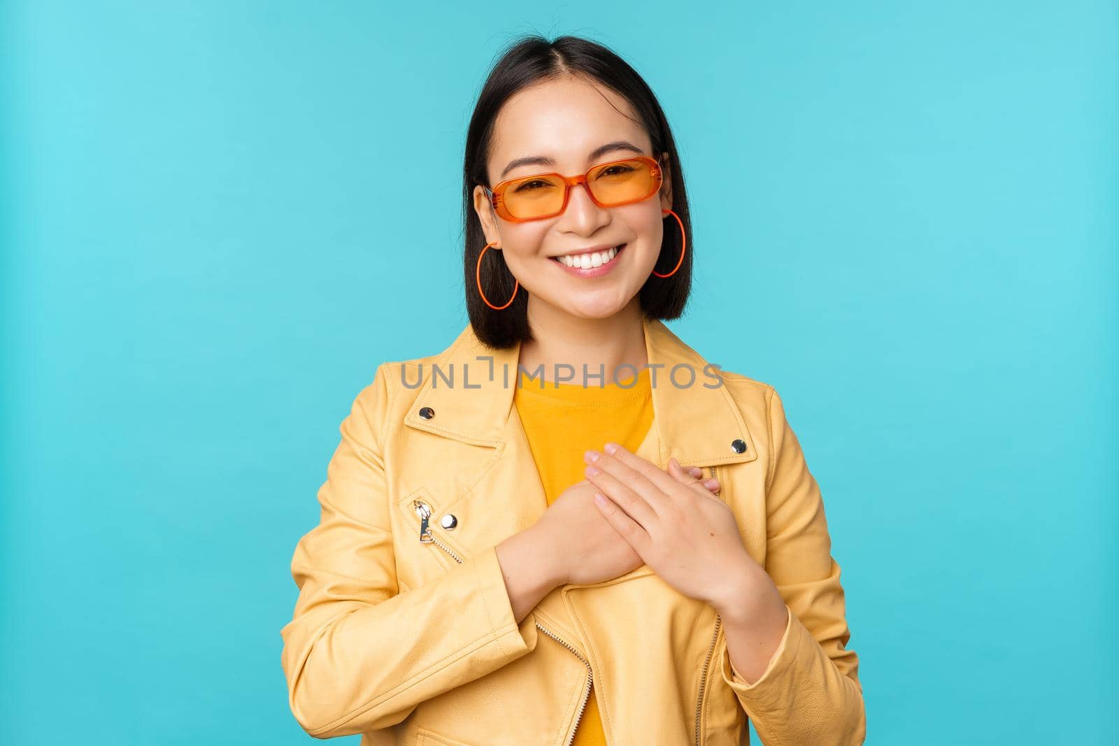 Portrait of asian woman smiling, holding hands on heart and looking with tenderness, care at camera, thankful emotion, standing over blue background by Benzoix