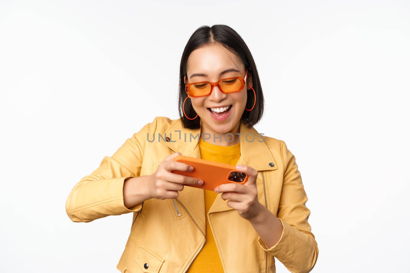 Stylish korean girl in sunglasses, playing mobile video game, laughing and smiling while using smartphone, standing over white background.