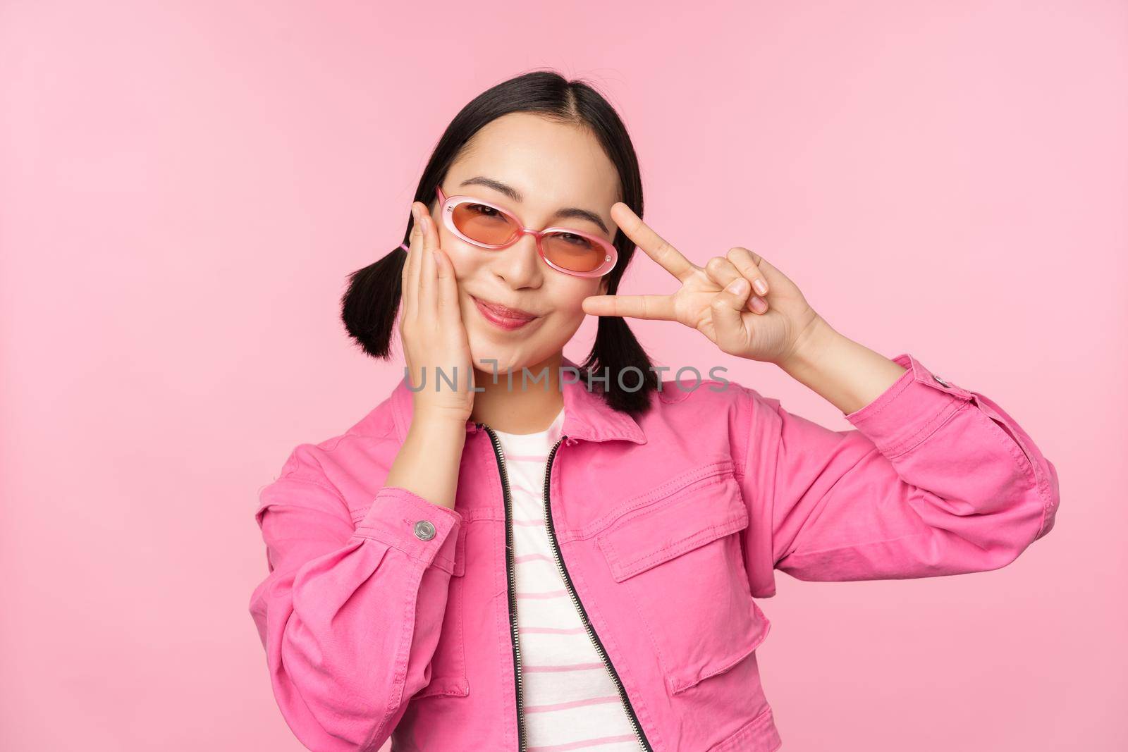 Portrait of kawaii asian girl in sunglasses, showing peace, v-sign near eye and smiles cute at camera, posing in trendy clothes against pink background by Benzoix