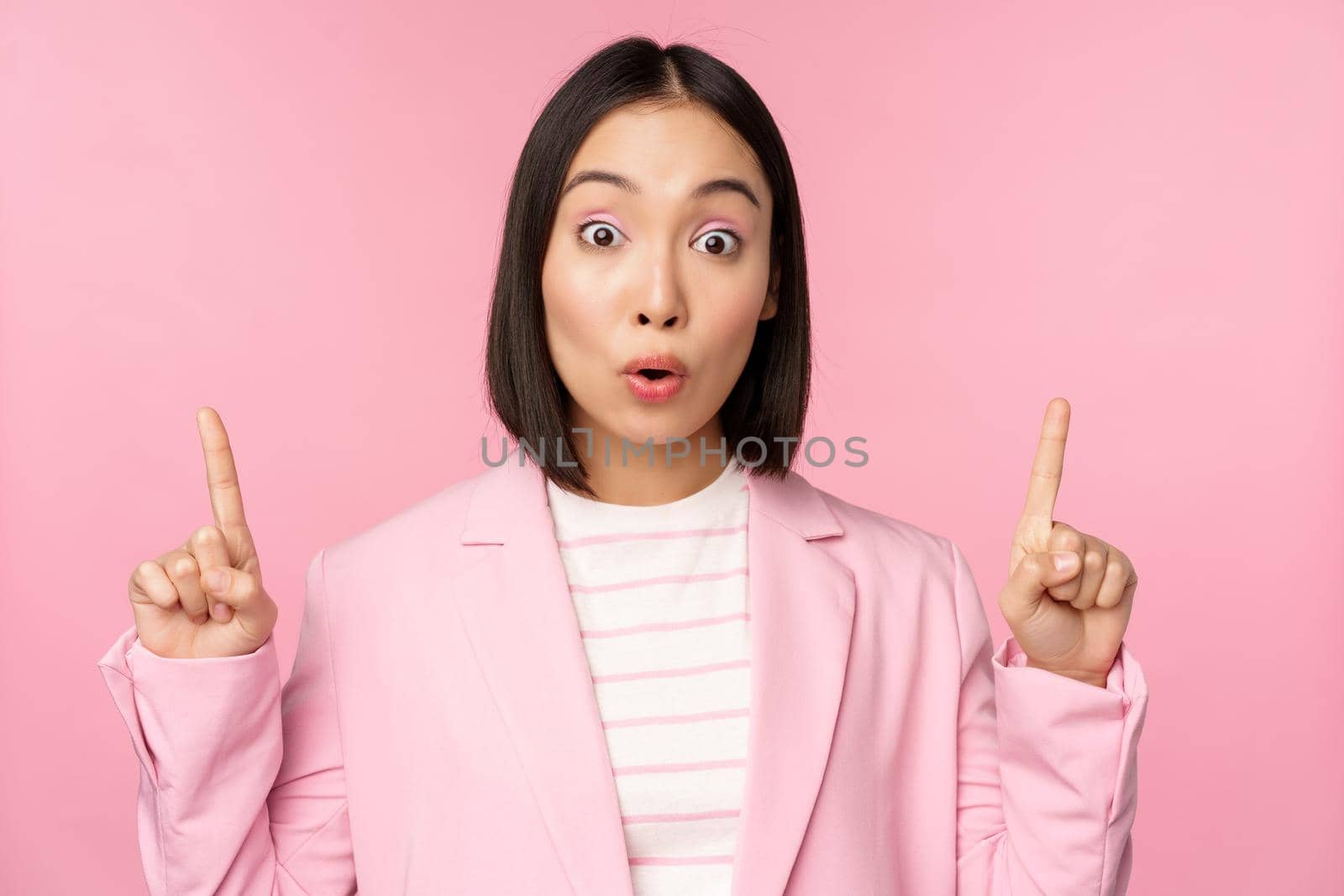 Portrait of asian businesswoman, corporate worker in suit, looks surprised by advertisement, points finger up, showing banner or logo on top, stands over pink background.