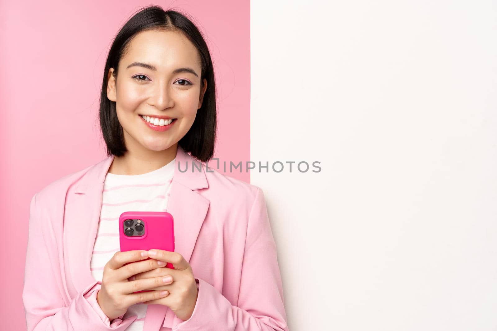 Image of korean female entrepreneur in suit, standing near info wall, advertisement on board, holding smartphone and smiling, posing over pink background.