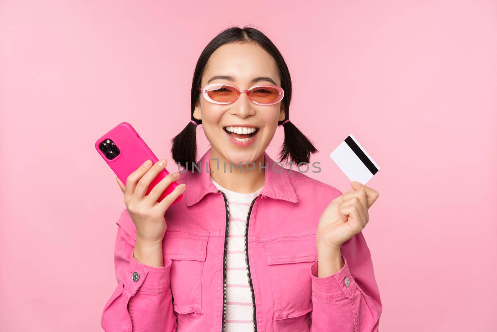 Modern beautiful asian girl, laughing and smiling with mobile phone, credit card, shopping online, paying with smartphone, standing over pink background by Benzoix