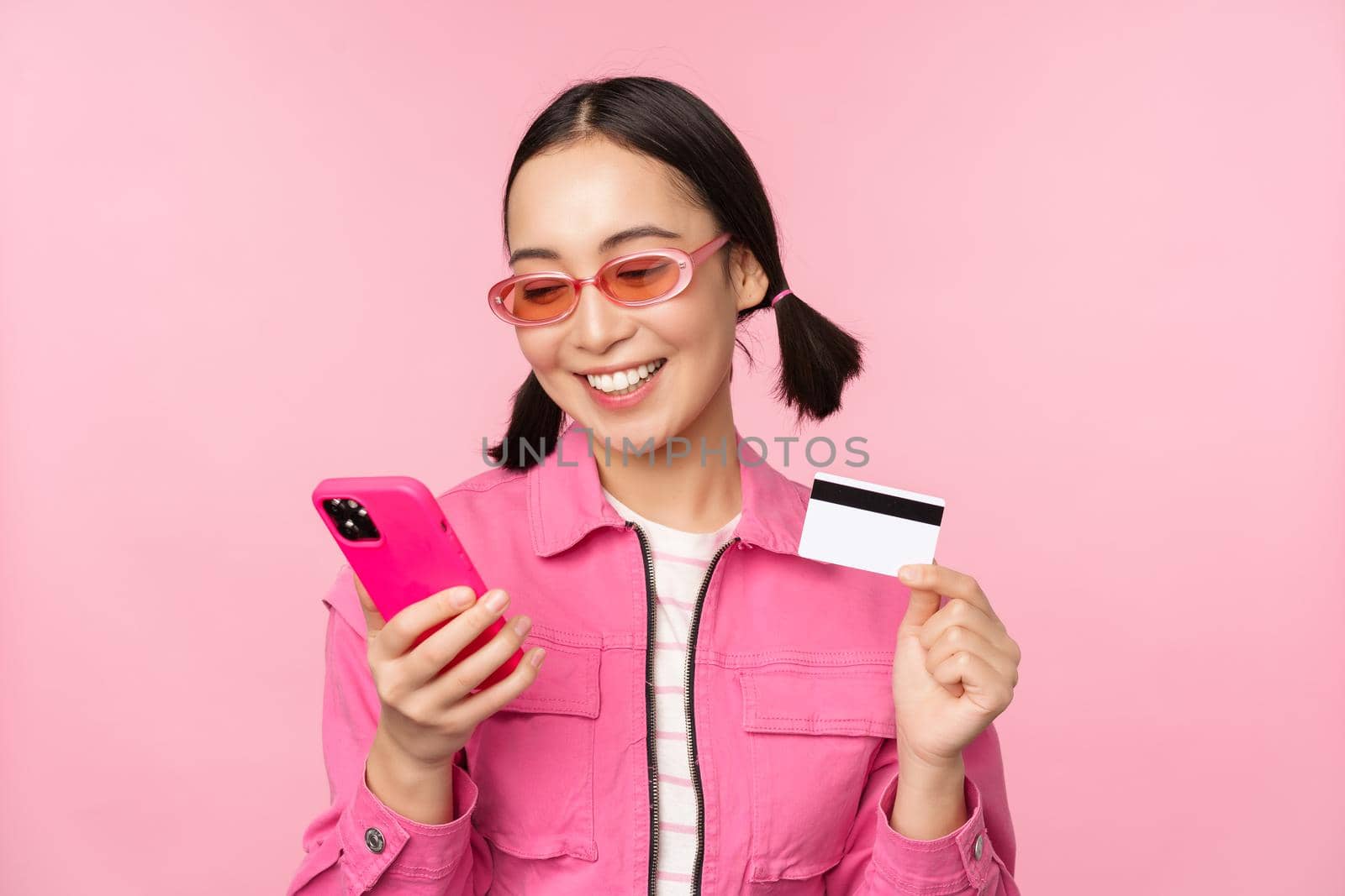 Online shopping. Smiling asian girl shopper, holding smartphone and credit card, paying in mobile app, standing over pink background.