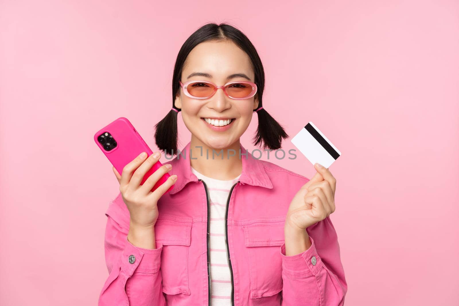 Beautiful korean woman holding smartphone, credit card, smiling at camera, buying online, shopping with mobile phone, standing over pink background.