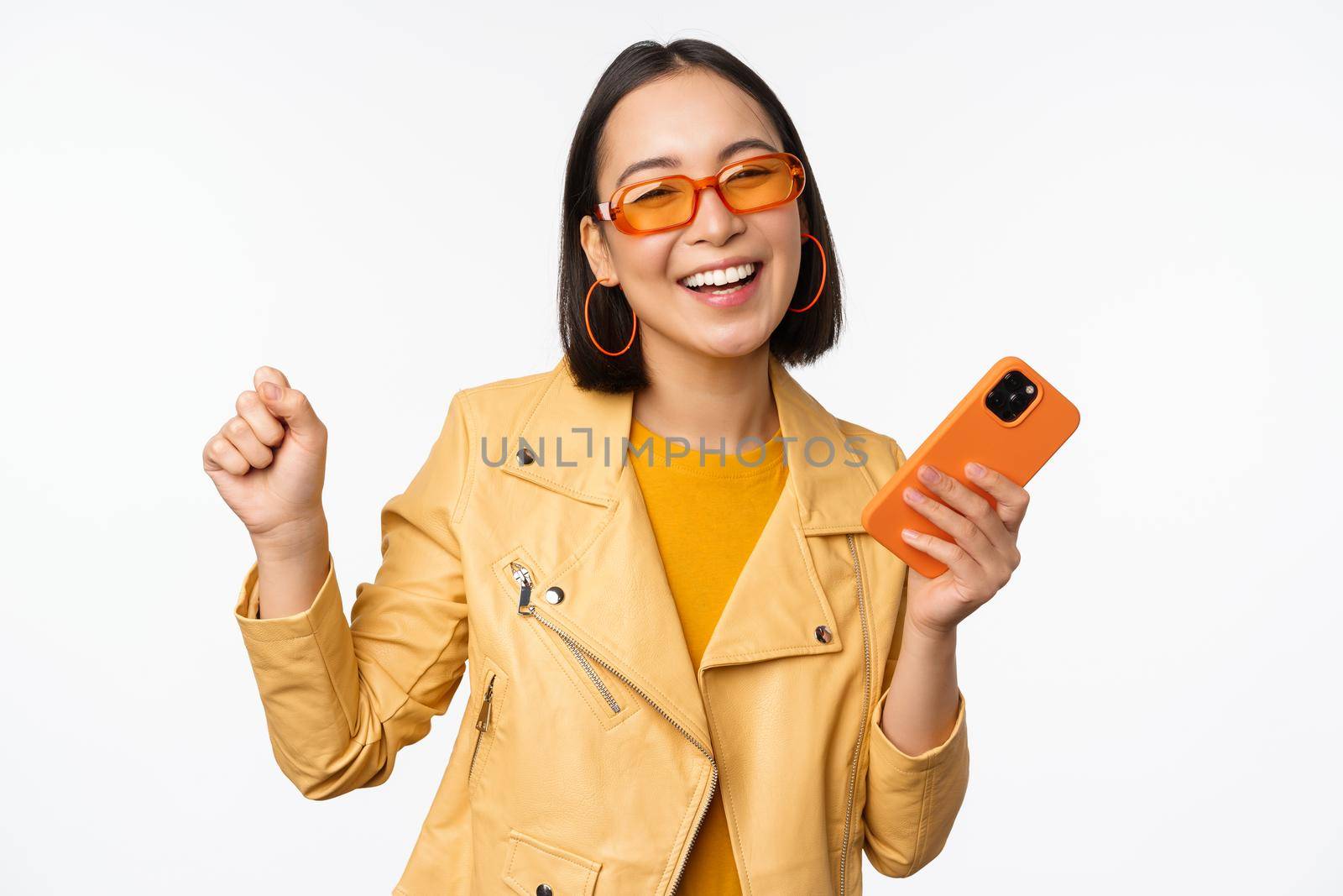 Image of stylish korean girl dancing with smartphone, laughing happy and smiling, standing over white background by Benzoix