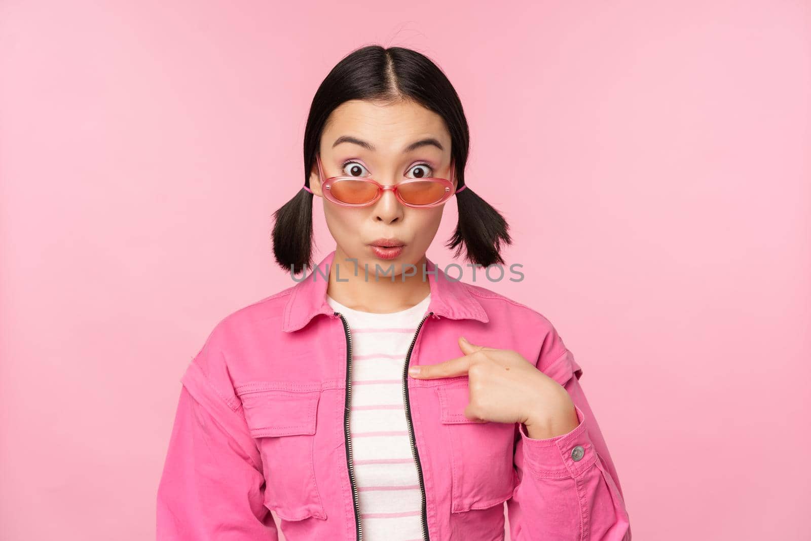 Close up portrait of asian girl looks surprised, points at herself with disbelief, being chosen, stands over pink background by Benzoix