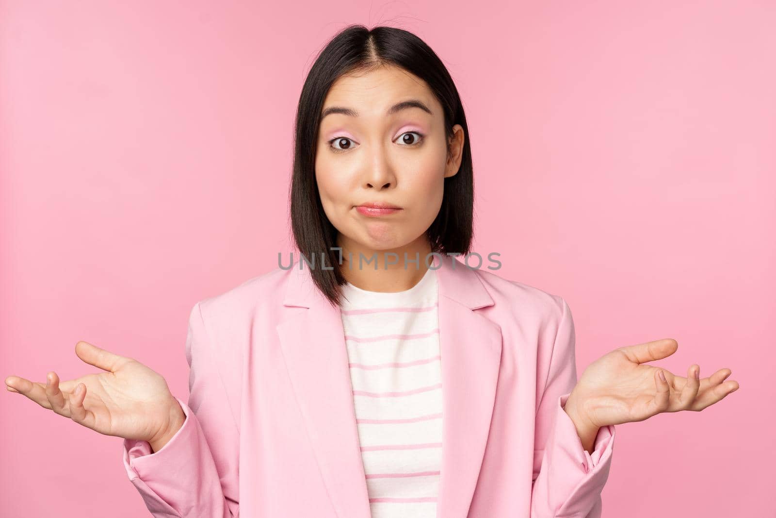 Portrait of confused asian businesswoman shrugging shoulders, looking clueless and puzzled, dont know, cant say, standing over pink background in office suit by Benzoix