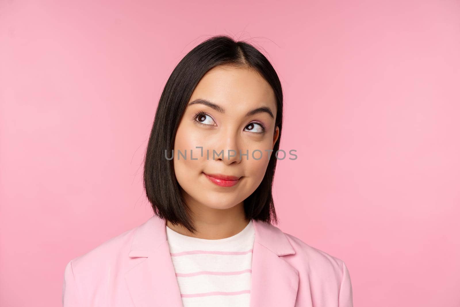 Close up portrait of young asian businesswoman thinking, smiling thoughtful and looking at upper left corner, standing over pink background by Benzoix