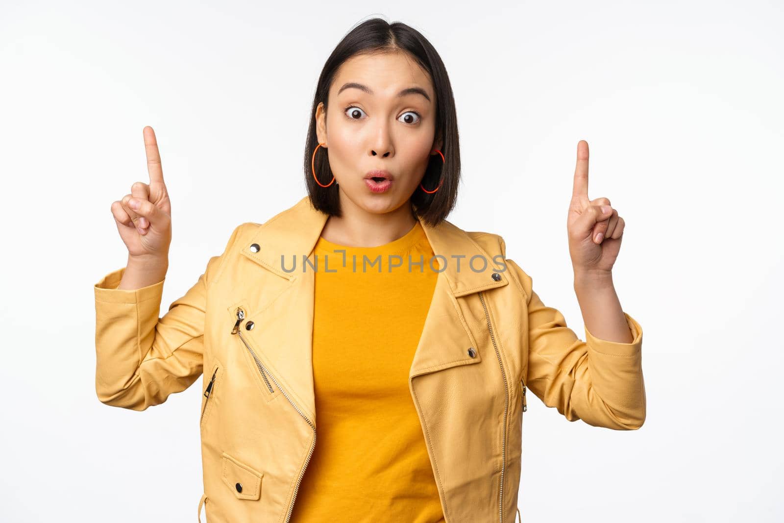 Surprised asian girl pointing fingers up, express interest, showing advertisement ahead, demonstrating promo banner on top, standing against white background.