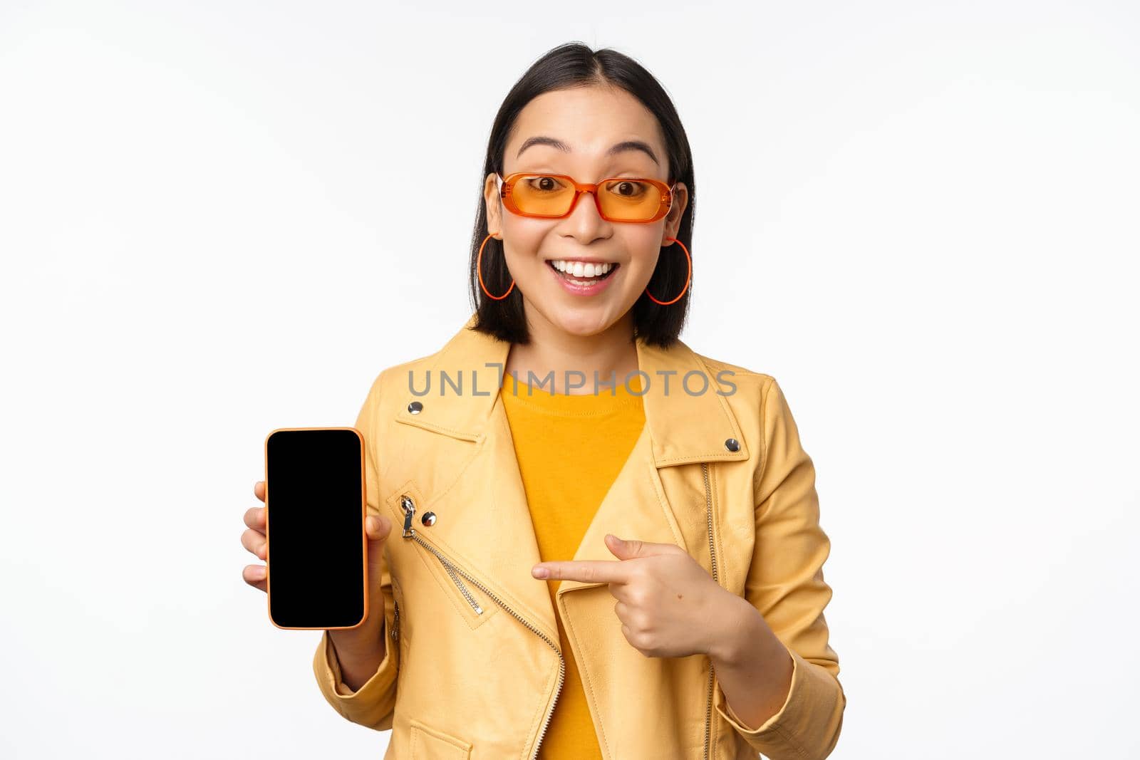 Portrait of stylish korean girl in sunglasses, smiling, pointing finger at smartphone screen, showing mobile phone application, standing over white background.