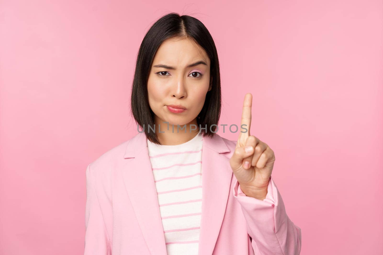 Asian businesswoman with serious, concerned face expression, showing stop motion, taboo, prohibit gesture, disapprove smth bad, standing over pink background.
