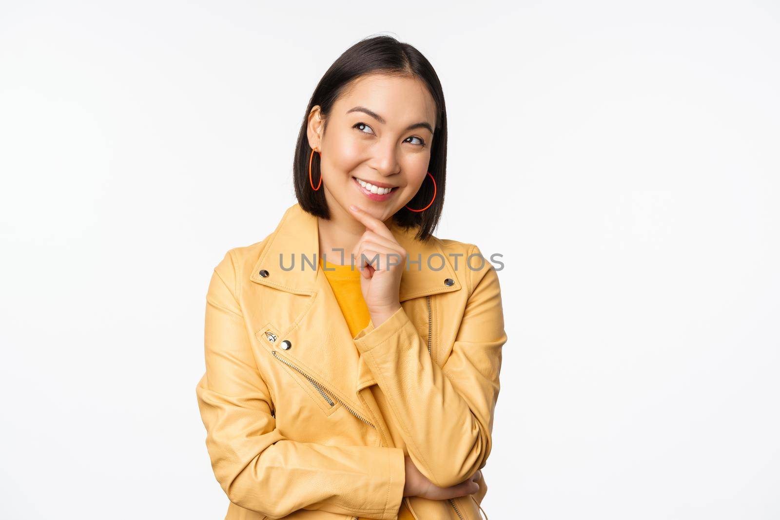 Portrait of stylish korean woman in yellow jacket, smiling thoughtful, thinking and looking up at logo or advertisement, standing over white background.