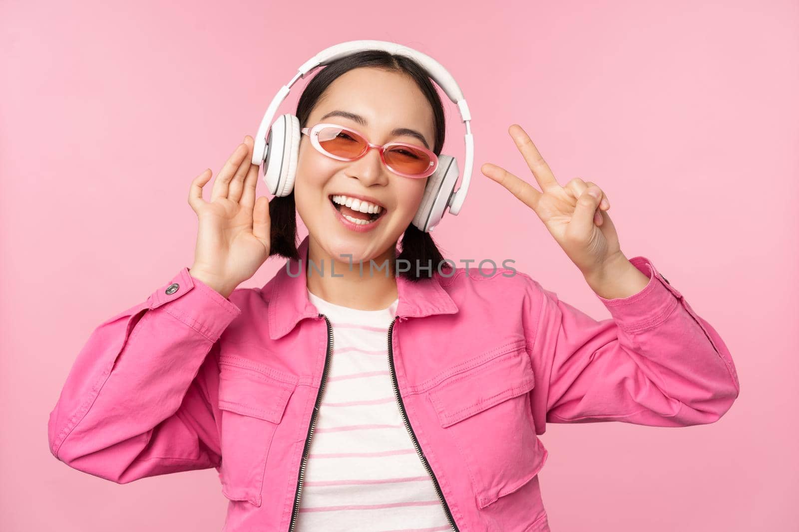 Dancing stylish asian girl listening music in headphones, posing against pink background.