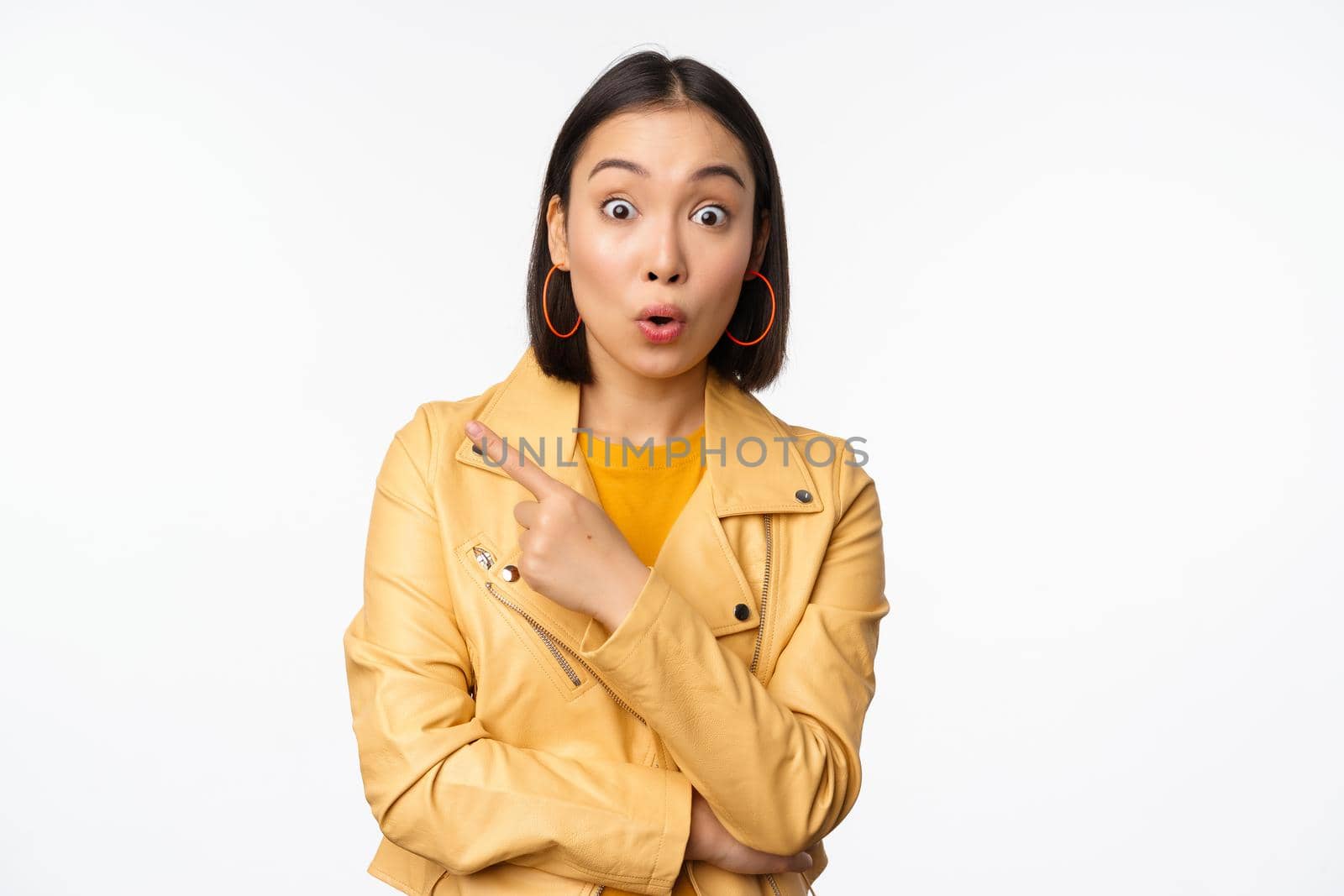 Portrait of beautiful asian woman express intereest and excitement about advertisement, store discount, pointing finger left, looking amazed, standing over white background by Benzoix