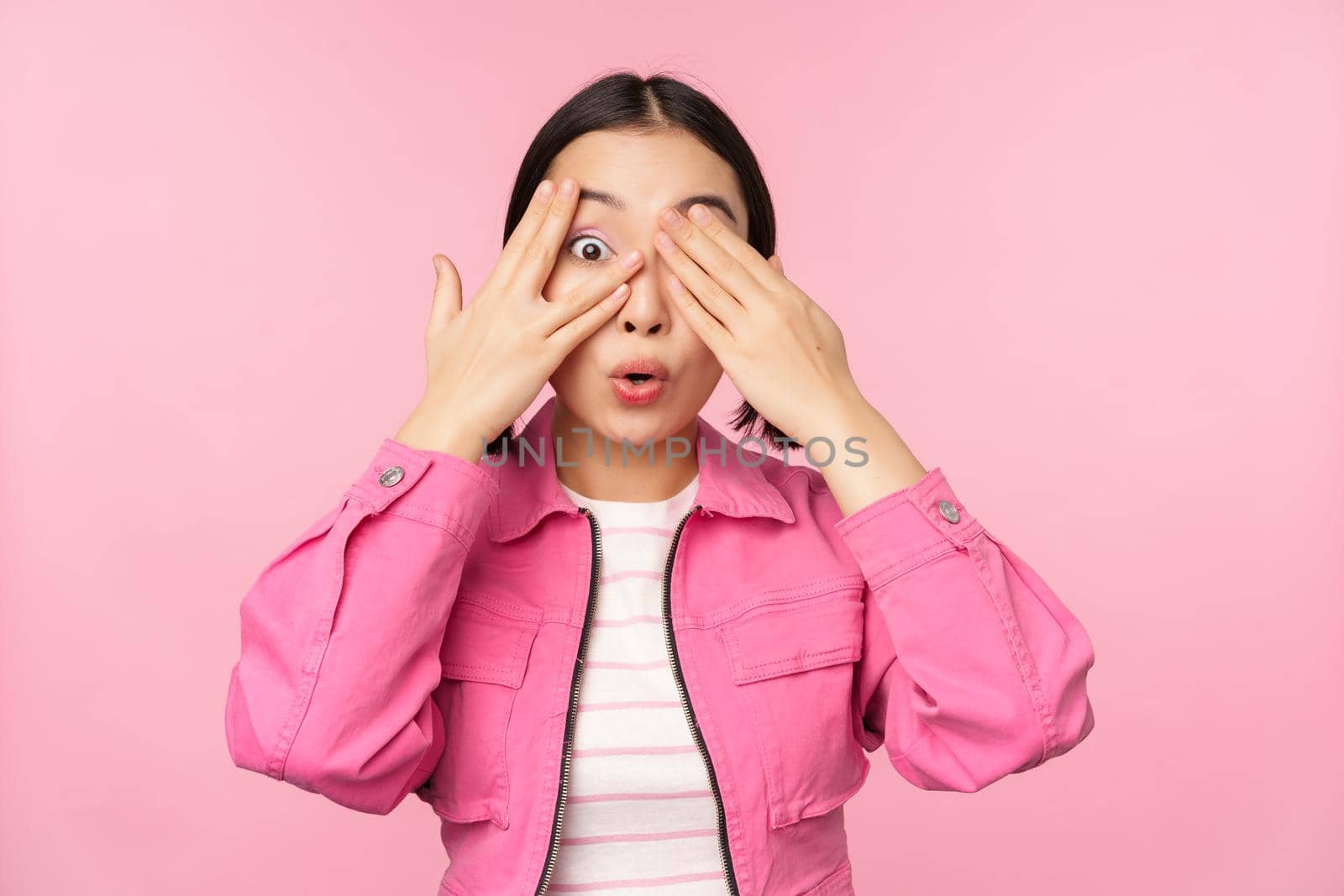 Portrait of asian girl peeks with excitement through fingers, covers eyes, seeing surprise, standing over pink background. Copy space