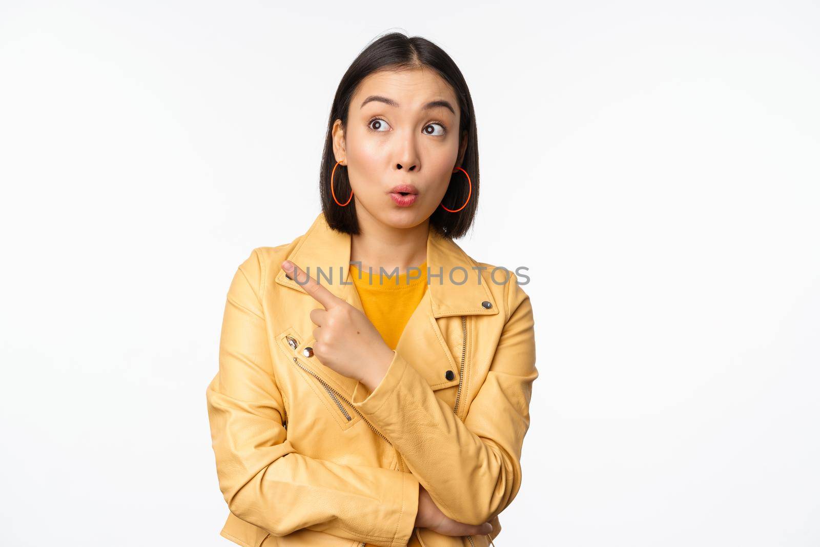 Image of surprised aisan woman pointing finger left, looking at logo or banner with intrigued face expression, standing over white background.