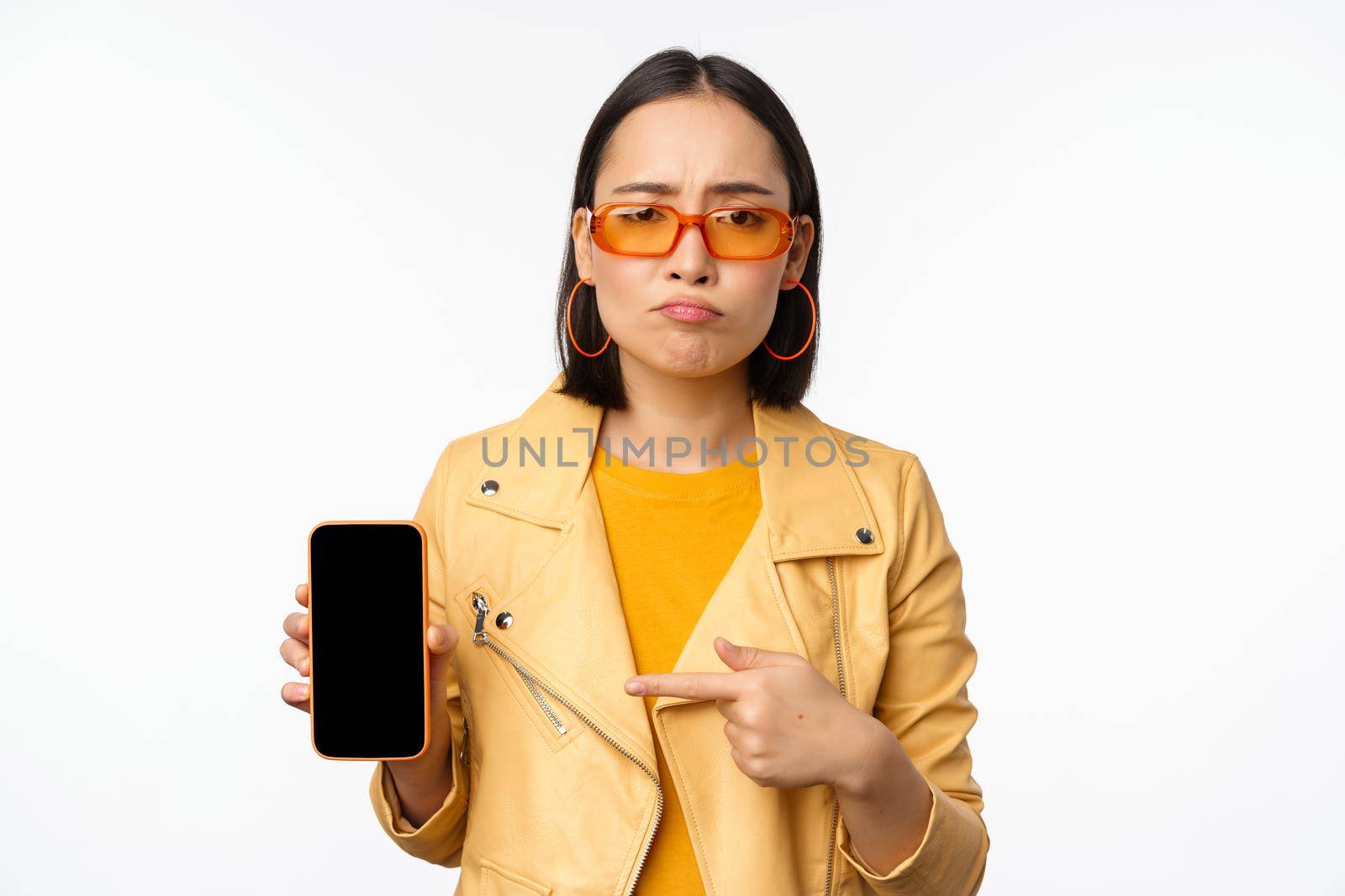 Portrait of sad asian woman in sunglasses, pointing finger at mobile phone app interface, showing smartphone application, standing over white background.