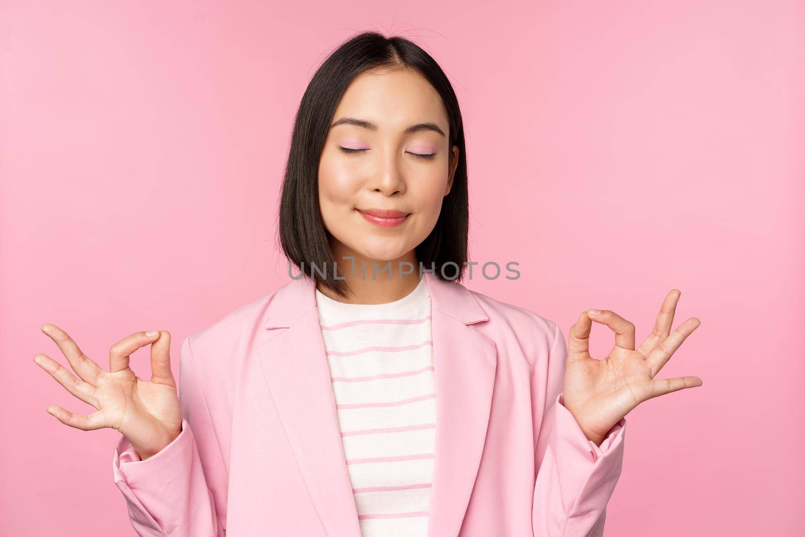 Keep calm. Smiling young asian woman meditating, practice yoga, mindfulness and relaxation at work, breath, inhale air with pleased face, standing over pink background by Benzoix