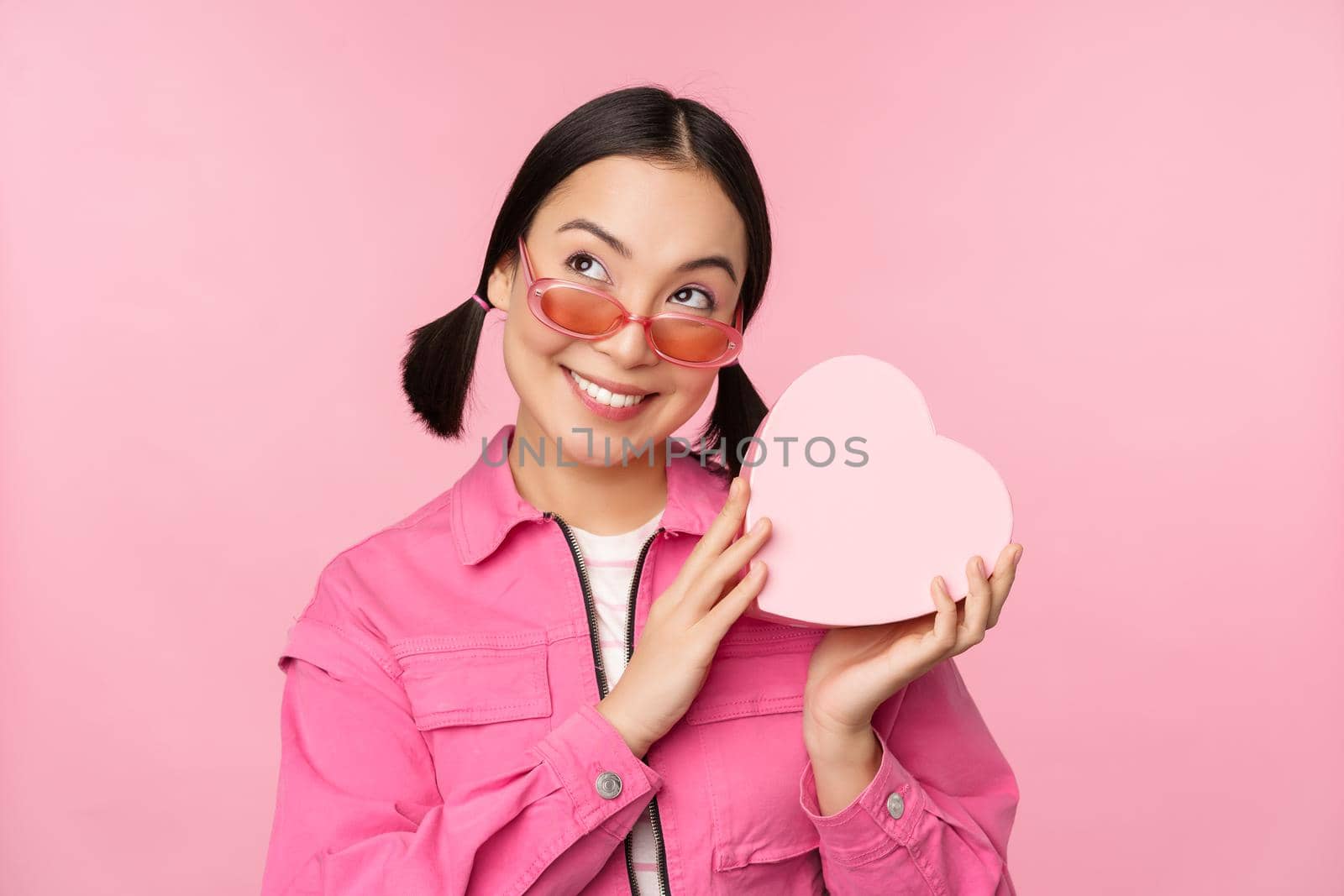 Image of stylish asian girlfriend in sunglasses, guessing whats inside gift box, heart-shaped present, standing over pink background.