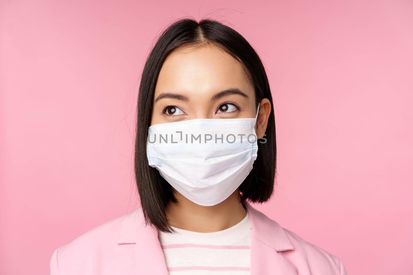 Close up portrait of japanese corporate woman in medical face mask from covid-19, looking left at logo, sale promo, standing over pink background.