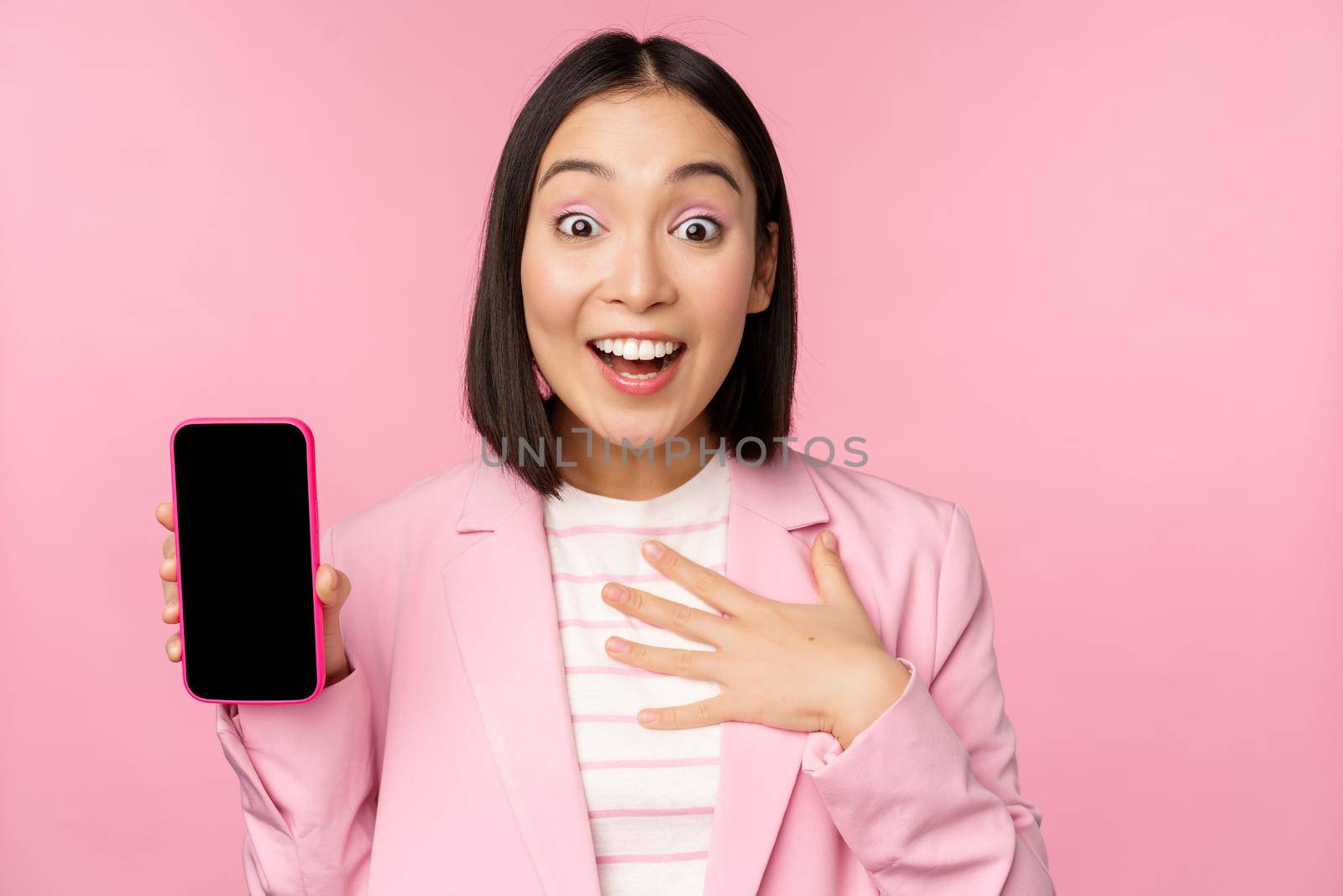 Surprised, enthusiastic asian businesswoman showing mobile phone screen, smartphone app interface, standing against pink background.