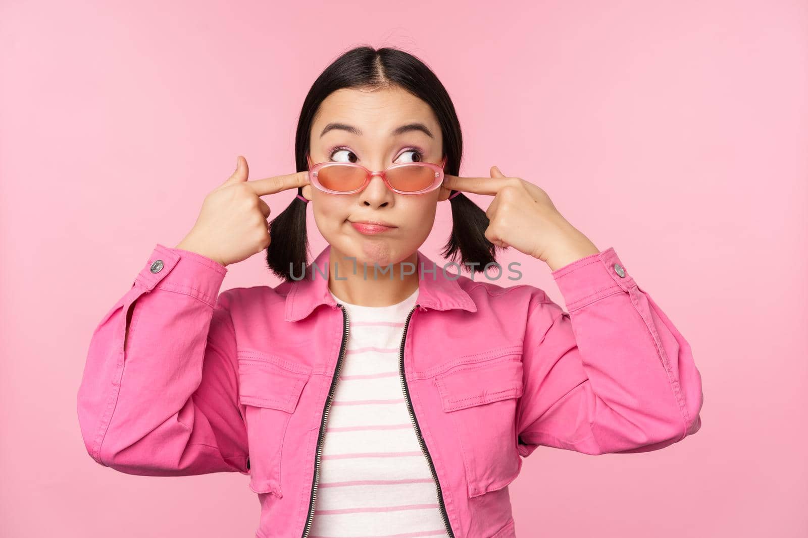 Portrait of silly japanese girl shuts ears, looks aside, doesnt listen, cant hear you gesture, standing over pink background, ignoring you by Benzoix