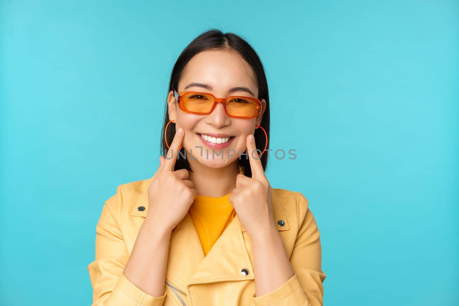 Close up portrait of asian young woman in sunglasses, smiling and looking romantic, standing happy over blue background by Benzoix