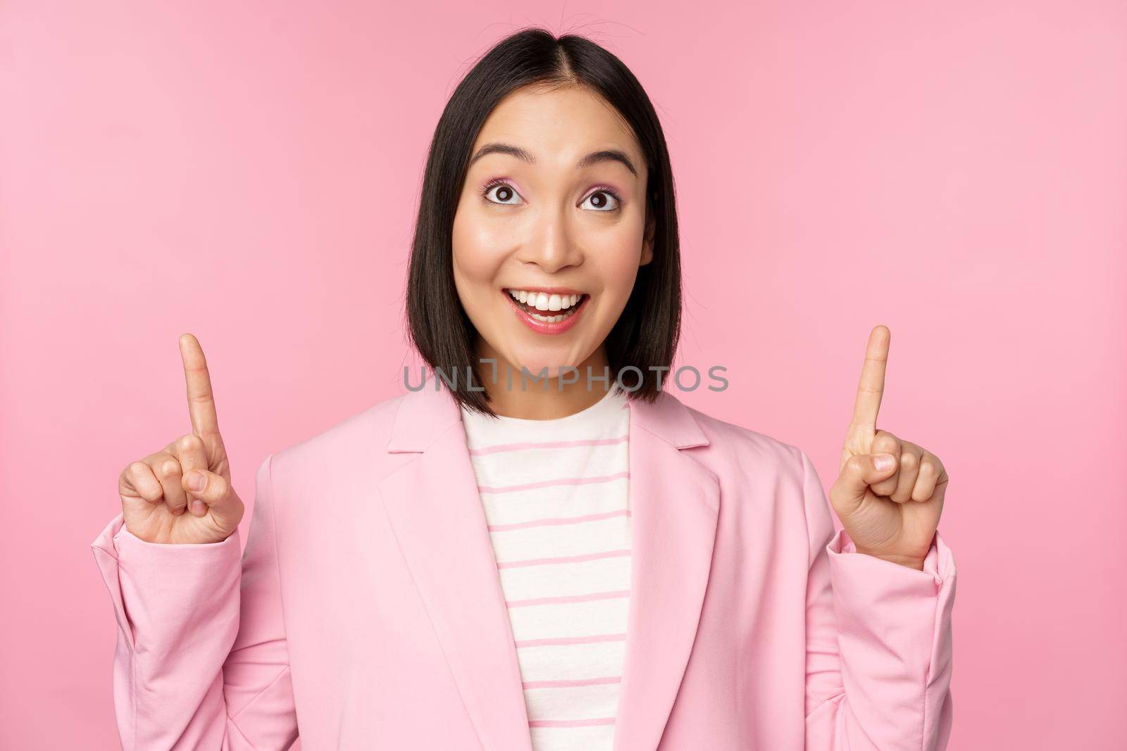 Enthusiastic corporate worker, asian business woman pointing fingers up and smiling, showing advertisement, logo, standing over pink background by Benzoix