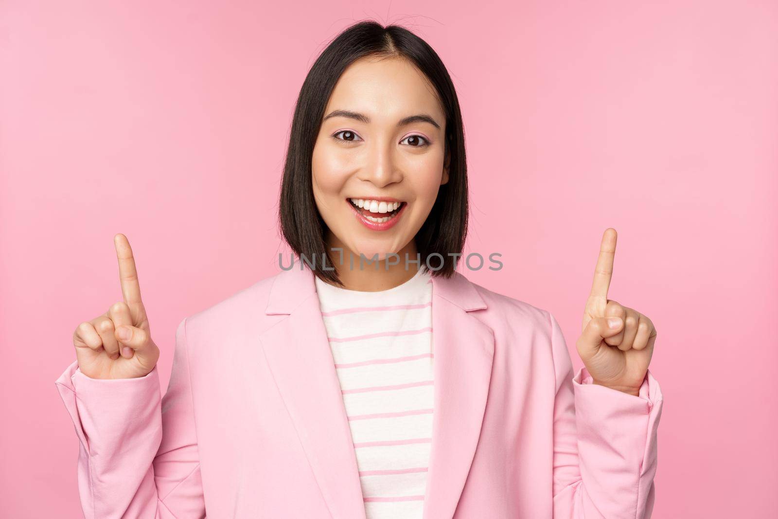 Smiling korean businesswoman, pointing fingers up, showing advertisement, banner or logo on top, standing in suit over pink background.