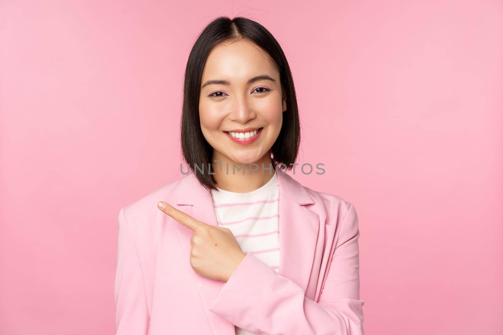 Portrait of asian business woman, saleswoman in suit pointing finger left, showing banner advertisement, smiling and looking professional, pink background by Benzoix