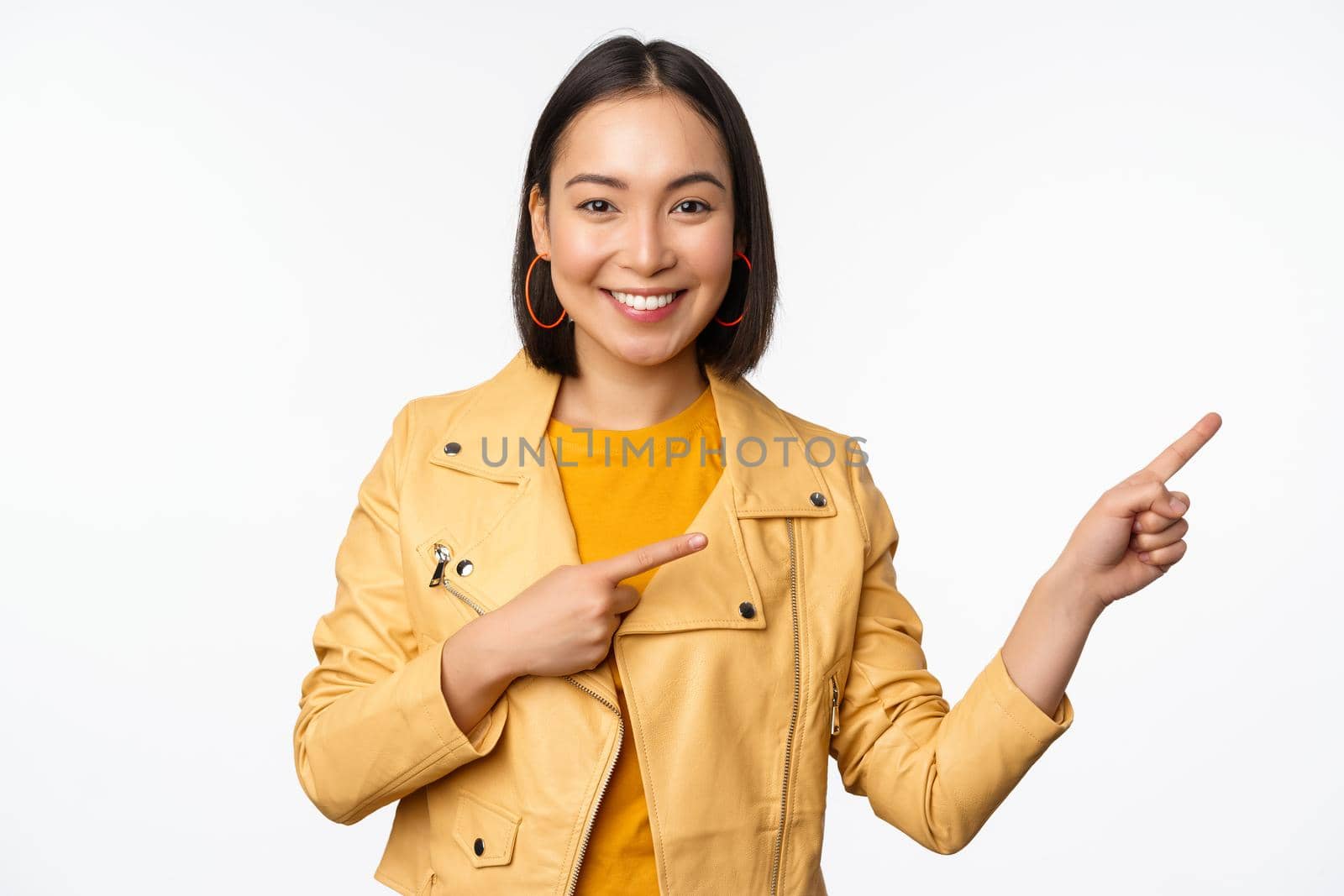 Happy asian woman smiling, pointing fingers right, inviting to check out sale, showing advertisement banner or logo, standing over white background.