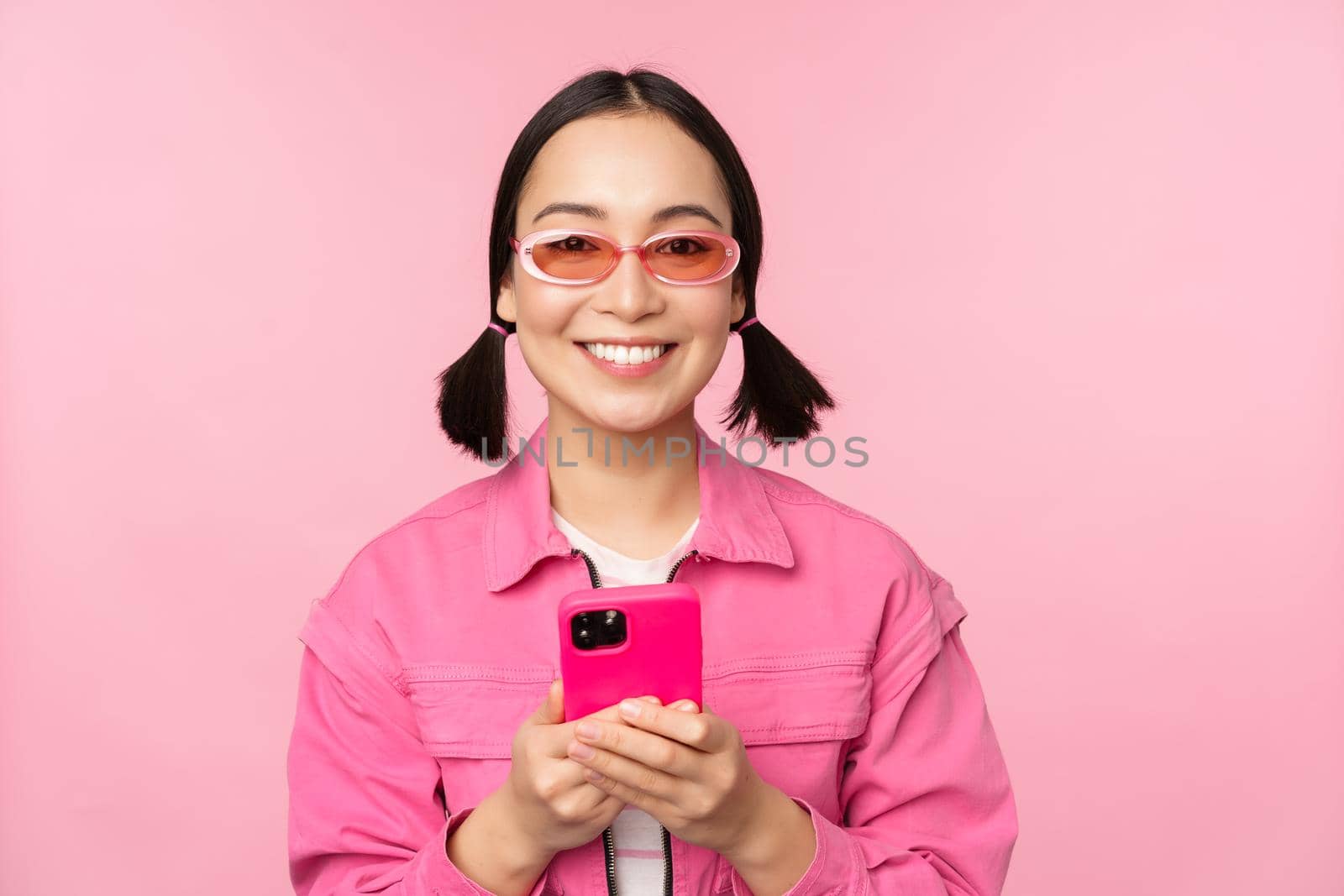 Portrait of korean girl in stylish sunglasses holding mobile phone, using smartphone app, standing over pink background. Copy space